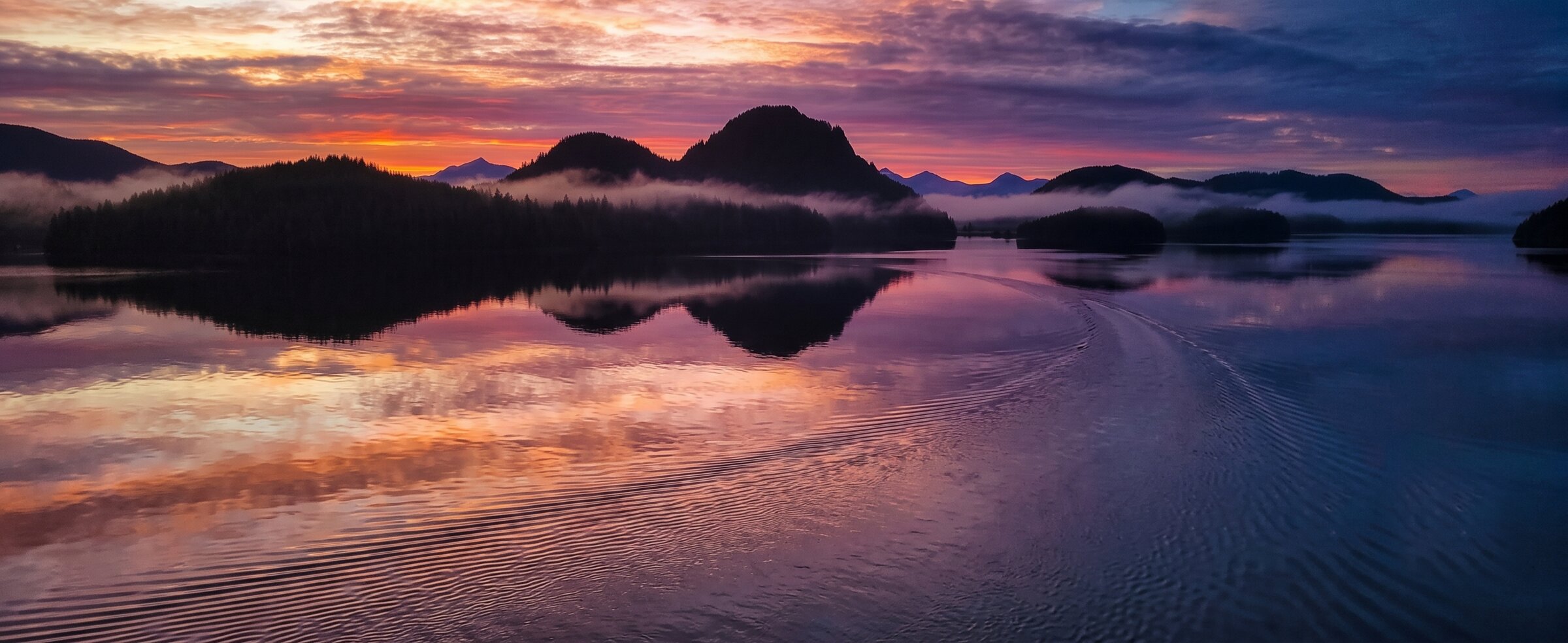 Vivid pink and orange sunset over calm water and misty islands on a beautiful evening sunset cruise near Tofino, British Columbia